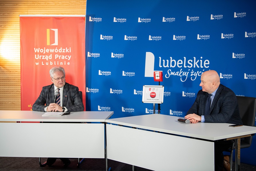 Zdjecie przedstawia dwóch męzczyzn siedzących przy stole konferencyjnym i rozmawiajacych. Za nimi dwie scianki z napisem lubelskie oraz wojewódzki urząd pracy w lublinie