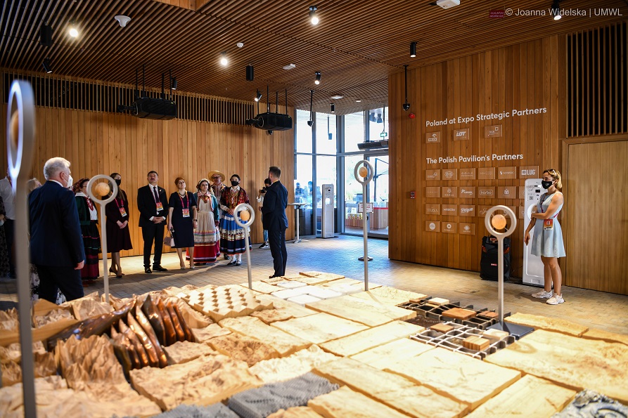 Zdjęcie przedstawia ludzi stojących w hali na wystawie, m.in znajdują się na zdjęciu tancerze z zespołu ludowego, stojący pod jedną ze ścian