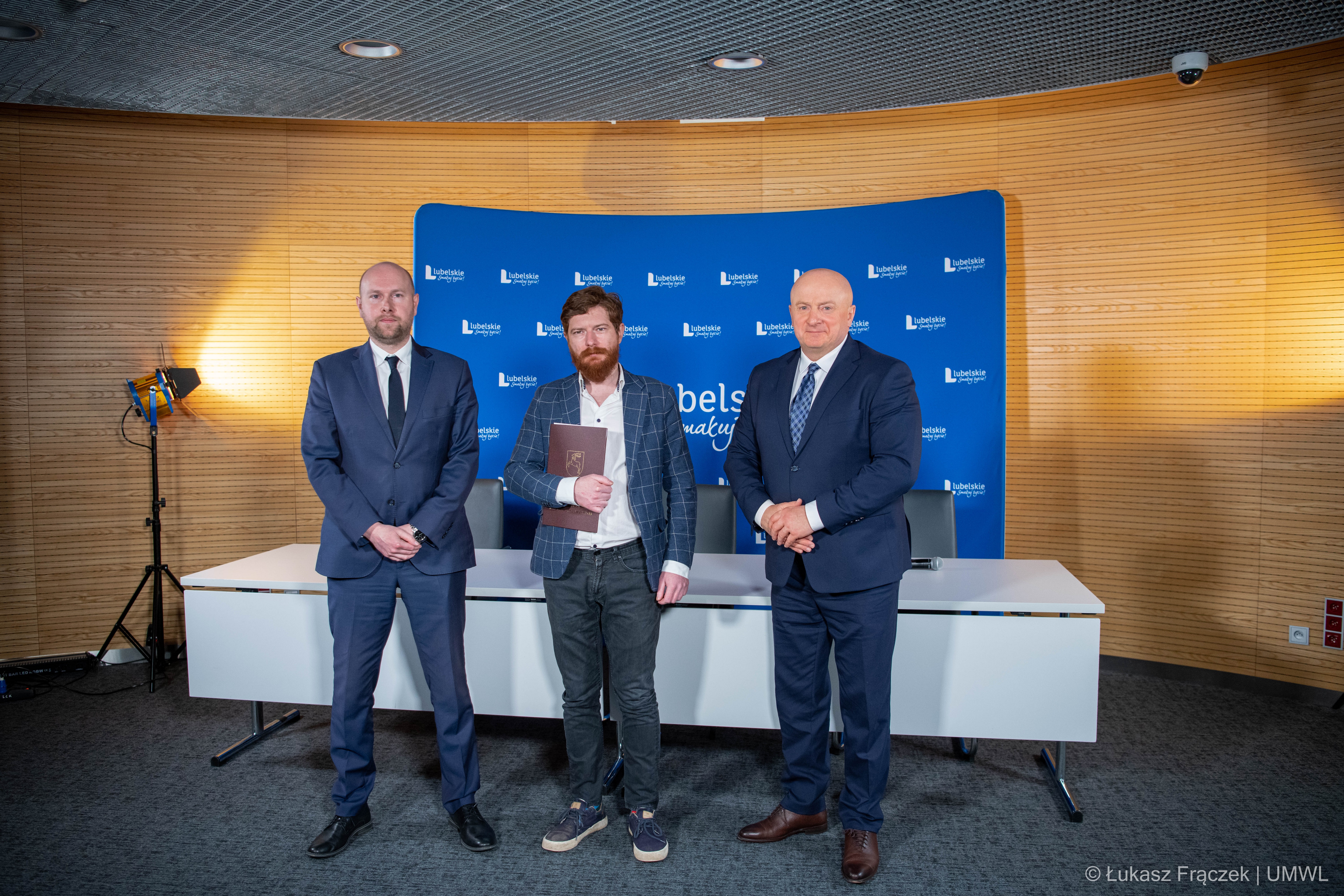 Zdjęcie przedstawia trzech mężczyzn stojących do zdjęcia w małej sali konferencyjnej, ten stojący w środku trzyma w rece teczkę na dokumenty