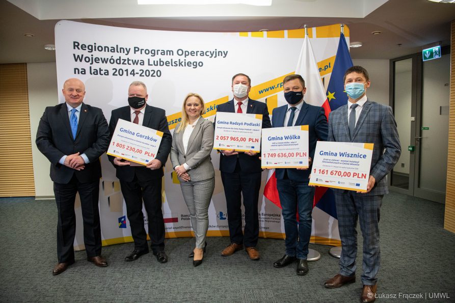 Zdjecie przedstawia grupe osób zarónow kobiet jak i mężczyzn którzy stoją obok siebie i trzymaja w dłoniach symboliczne umowy na dofinansowania