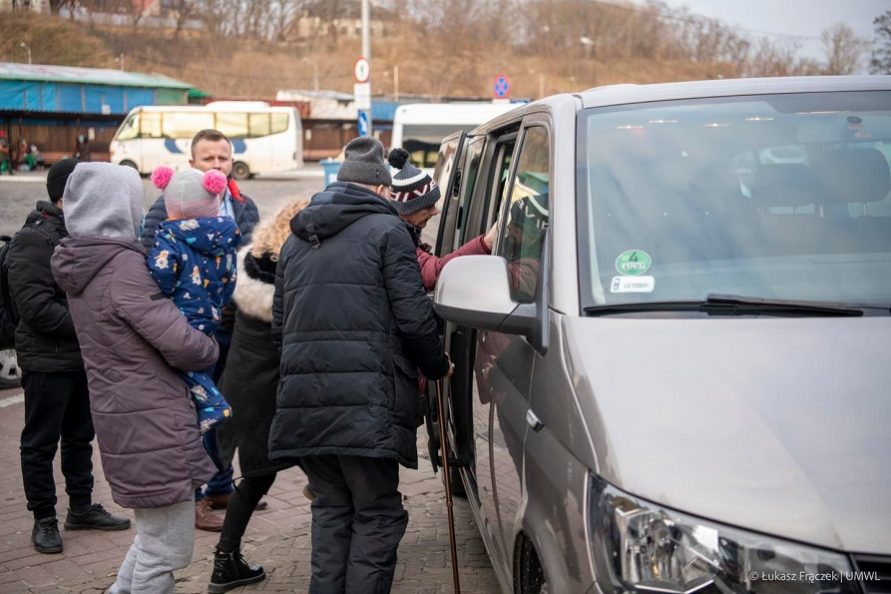 Zdjęcie przedstawia samochód, duży bus do którego wsiadaja osoby róznej płci i wieku