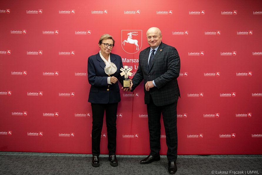 Na zdjeciu znajdują się dwie osoby kobieta i męzczyzna. Meżczynza przekazuje kobiecie nagrodę w postaci statuetki