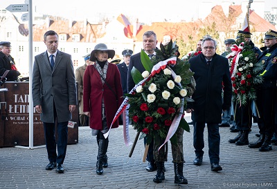 Zdjecie przedstawia 4 osoby trzech mężczyzn i kobietę, stojacych na dworze, jeden z mężczyzn niesie wieniec kwiatów