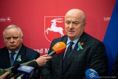 na zdjęciu widać dwóch mezczynz stojących wewnątrz pomieszczenia jeden z nich udziela wypowiedzi do mikrofonu