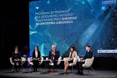 Na zdjęciu znajduje sie dwoje doroslych osób i troje uczniów, ktorzy siedza na fotelach na scenie w sali konferencyjnej