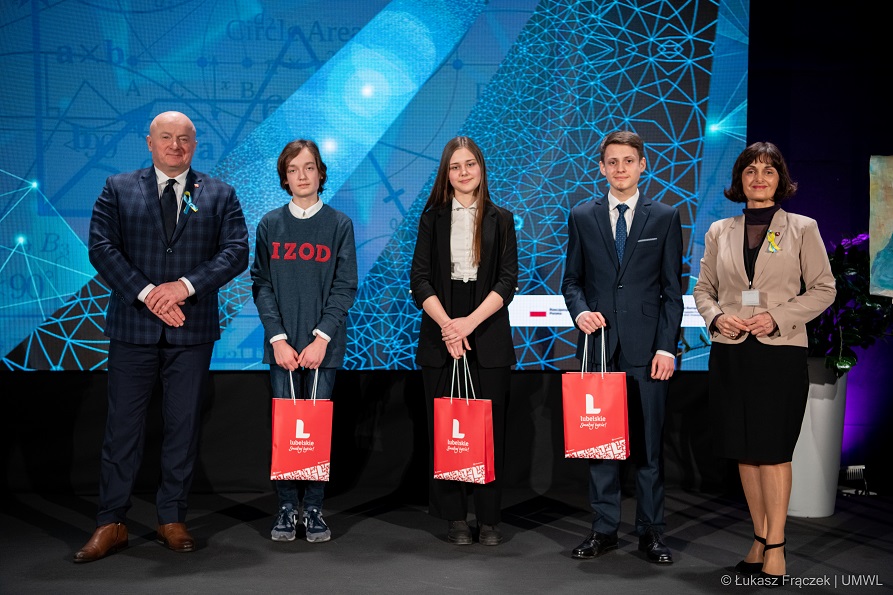 Na zdjęciu widać dwie stojące osoby dorosłe i troje uczniów, uczniowie trzymają w rękach torebki prezentowe