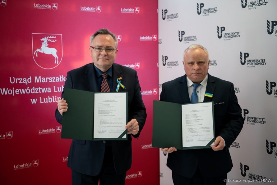 Zdjęcie przedstawia dwóch mężczyzn którzy stoją obok siebie i pozują do zdjęcia trzymając w rekach podpisane porozumienie