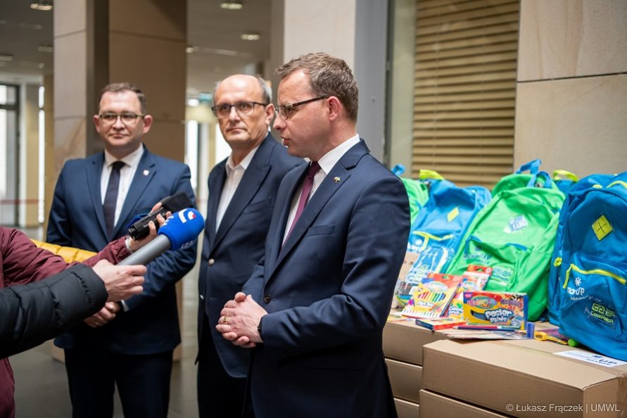 Trzech mężczyzn stojących na korytarzu, jeden z nich udziela wypowiedzi do mikrofonu, za nimi stoją przygotowane pakiety przyborów szkolnych do przekazania dzieciom z Ukrainy