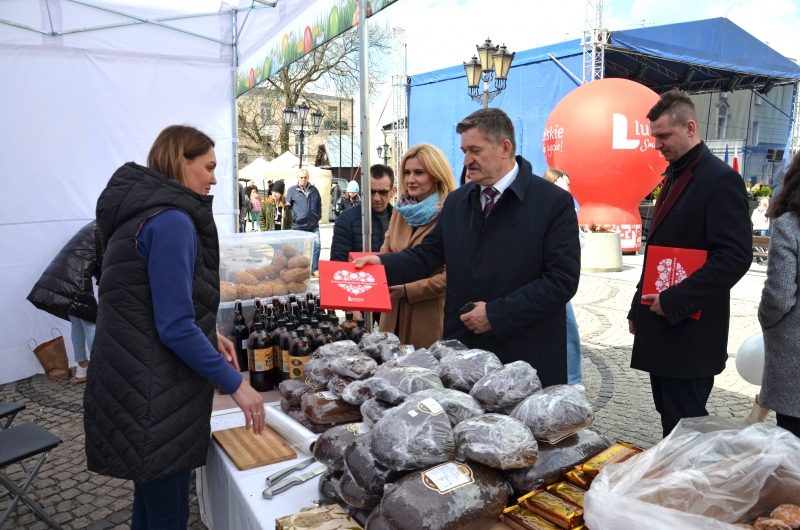 Mezczyzna i kobieta przekazuja książkę kobiecie, która stoi przy swoim stoisku z wyrobami podczas jarmarku
