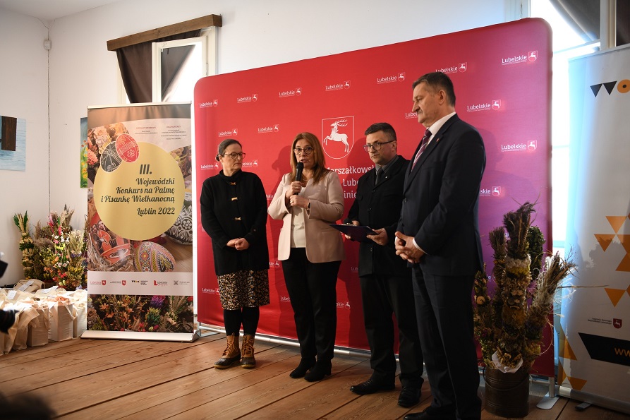 Dwie kobiety i dwóch meżczyzn stoją w sali,, jedna z kobiet mówi do mikrofinu. Za nimi stoją banery promujące województwo i odbywajacy się konkurs wielkanocny