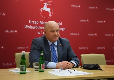 Mężczyzna w dojrzałym wieku siedzi przy stole w sali konferencyjnej. Na stole znajduje się mikrofon, butelki z wodą oraz notatki