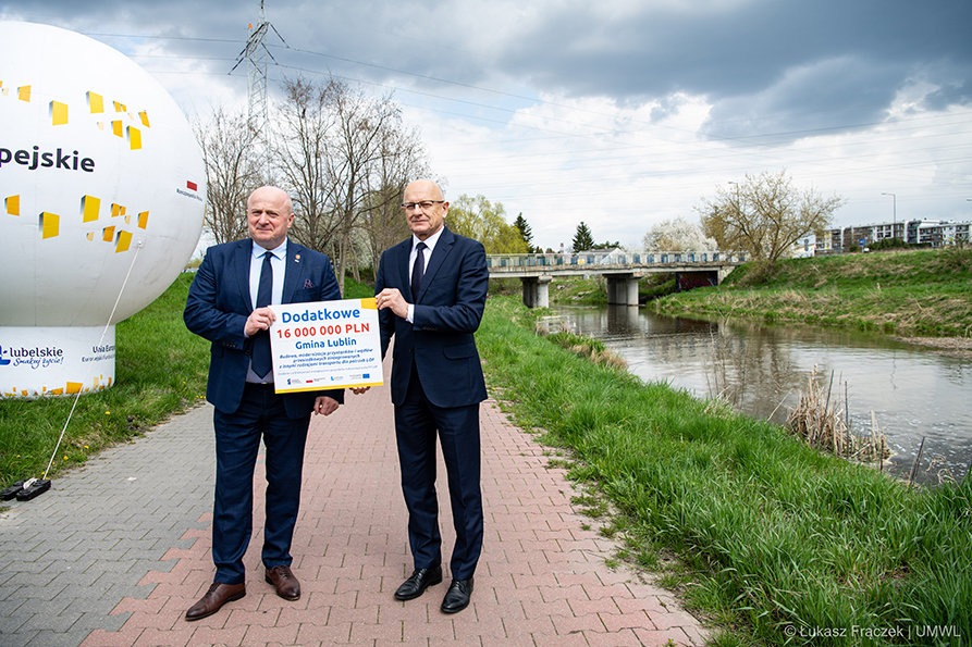 Dwóch mężczyzn stoi na chodniku i trzymąją symboliczną kartę z napisana kwotą dofinansowania. W tle za nimi znajduje się most i mała rzeka