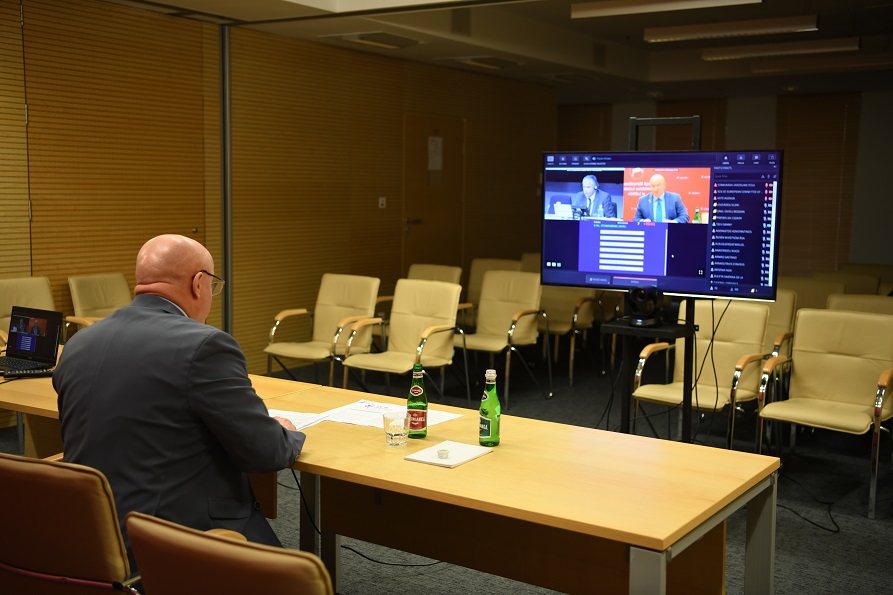 Widok na salę konferencyjną z ustawionym dużym ekranem na którym widać uczestników zdalnego spotkania. W sali plecami do aparatu siedzi mężczyzna