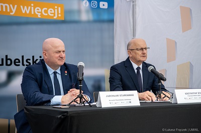 Dwóch mężczyzn w dojrzałym wieku siedzi przy stole postawionym w namiocie na dworze. Na stole stoją mikrofony i wizytowniki z ich imionami i nazwiskami.