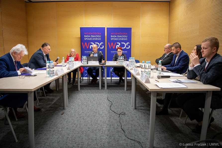 Grupa osób siedziących w sali konferencyjnej przy stołach ustawionych w podkowę