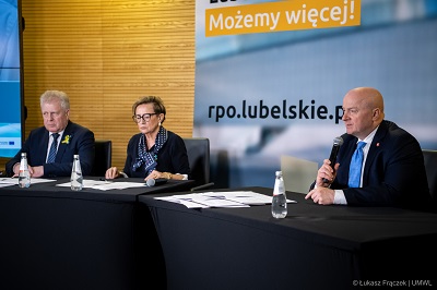 Dwóch mężyczn i jedna kobieta siedzacy przy stolikach w sali konferenacyjnej, jeden z mężczyzn udziela wypowiedzi