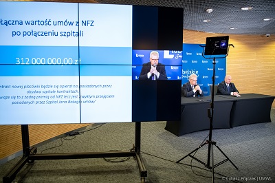 Widoczny na pierwszym planie telebim a zanim widok na dwóch meżczyzn siedzacych przy stole konferencyjnym