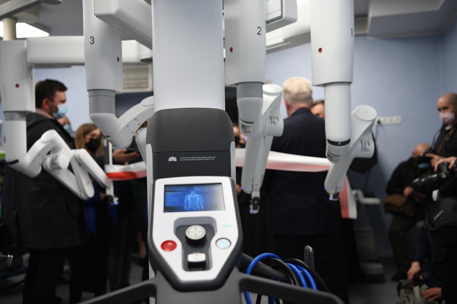 Na pierwszym planie nowoczesny robot operacyjny a w tle grupa ludzi przybyłych na otwarcie sali operacyjnej