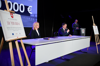 Przy podłużnym stole siedzi umieszczonym w sali konferencyjnej siedzi kobieta i mężczyzna. Mężczyzna trzyma mikrofon. Po przeciwległych końcach stołu znajdują się ustawione sztalugi z informacjami o wysokości dotacji.