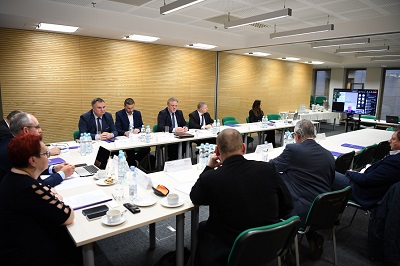 Grupa osób siedzących przy stołach ustawionych w podkowę w sali konferencyjnej