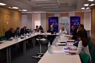 Grupa kobiet i mężczyzn siedzi przy stołach ustawionych w podkowę w sali konferencyjnej. Przed nimi na stołach leżą notatki i mikrofony.