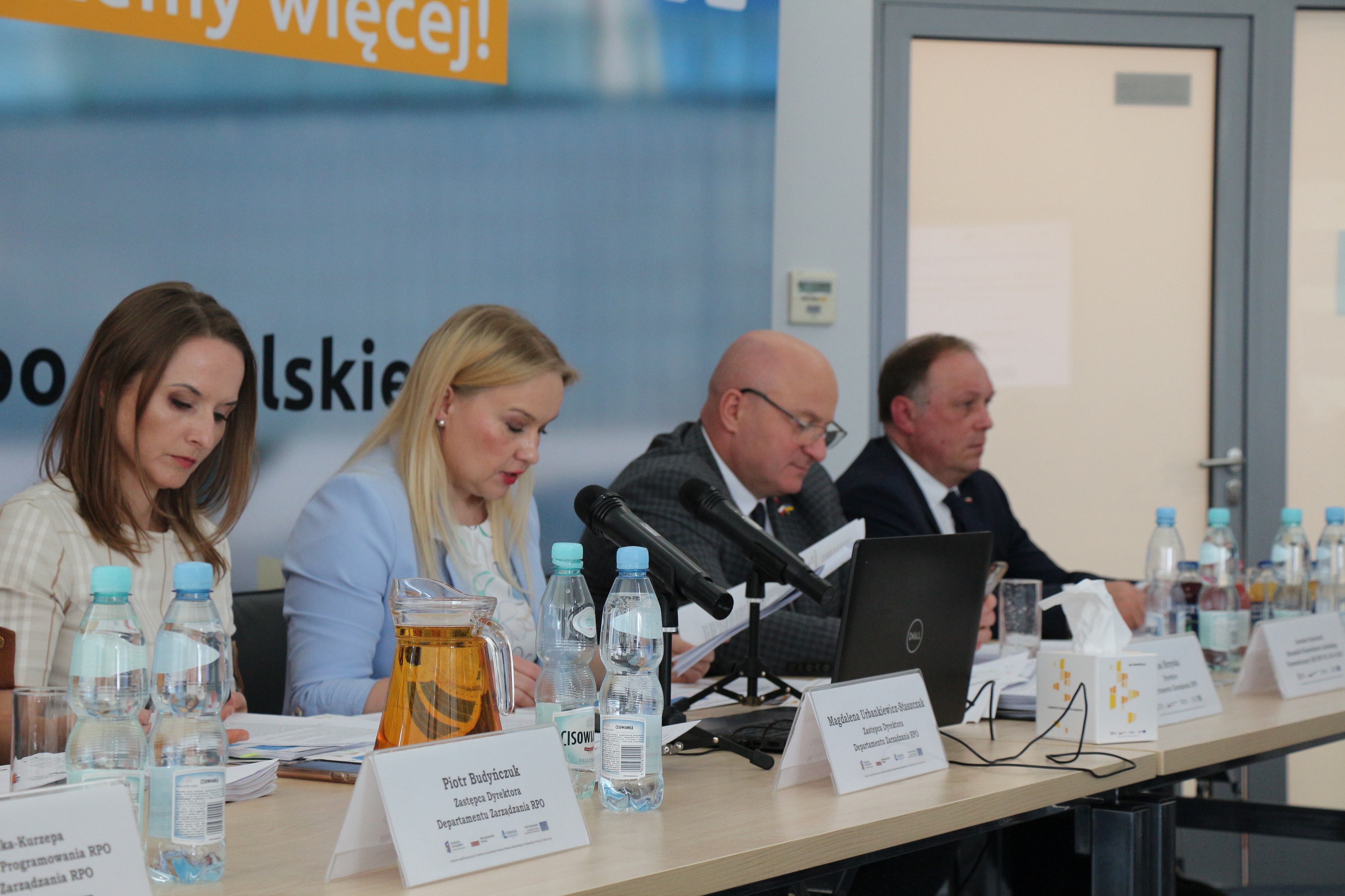 Zbliżenie na 4 osoby siedzące przy stole konferencyjnym. Kobieta siedząca najbliżej udziela wypowiedzi. Na stole znajdują się wizytowniki, woda butelkowana i notatki