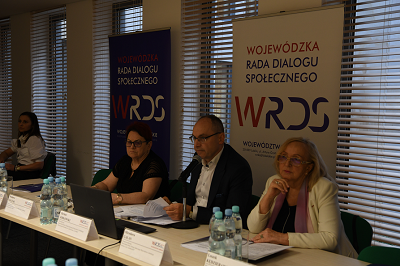 Dwie kobiety i mężczyzna siedzą przy stole w sali konferencyjnej. Przed nimi stoi butelkowana woda oraz leżą notatki i mikrofony. W tle widać ustawione banery znapisem WRDS Wojewódzka Rada Dialogu Społecznego