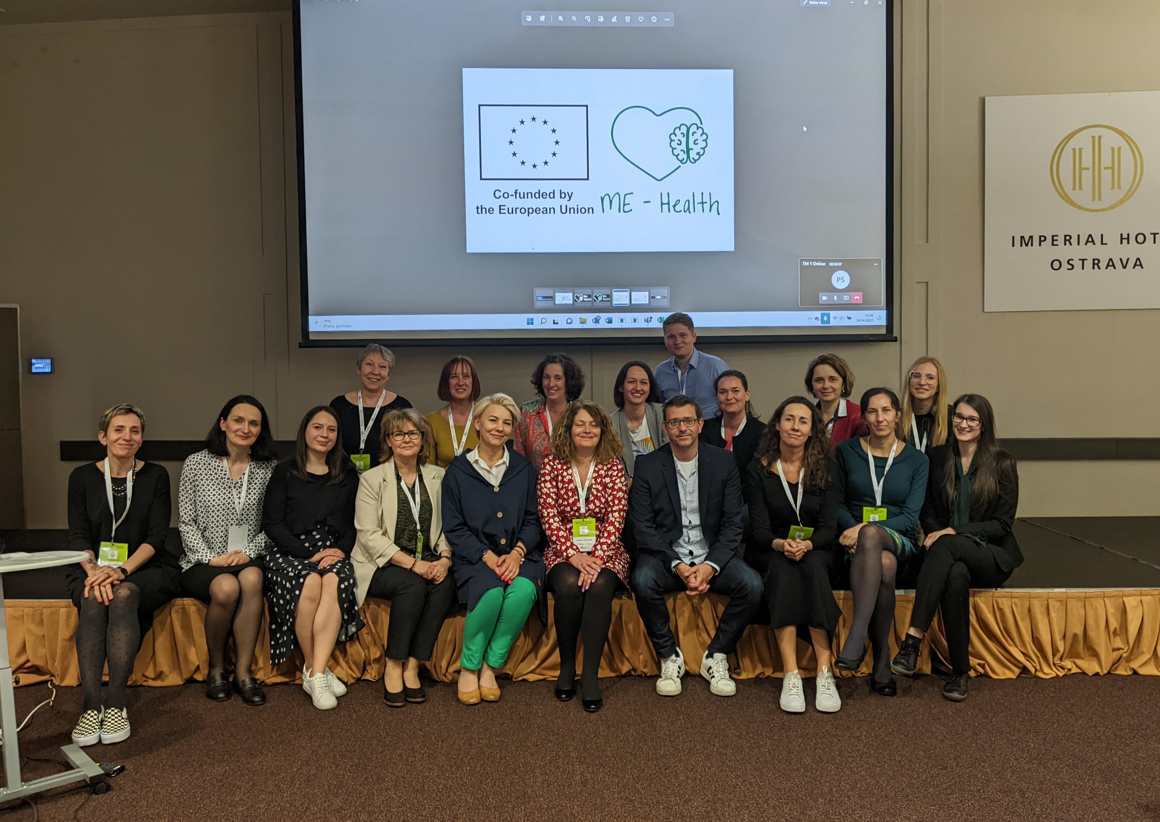 Zdjecie przedstawia liczną grupe osób, ustawioną w dwóch rzędach i pozujacą do zdjęcia. Za nimi w tle znajduje się ekran z wyświetloną prezentacją