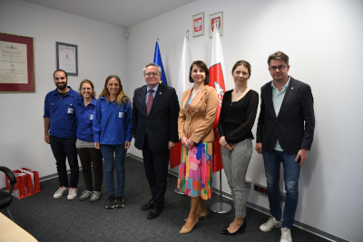 Grupa osób, kobiet i mężczyzn stoi w pomieszczeniu i pozują do zdjęcia. Za nimi znajdują się ustawione flagi