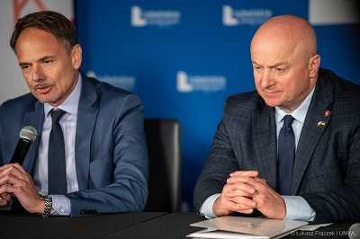 Dwóch mężczyzn siedzi przy stole w sali konferencyjnej, jeden z nich trzyma mikrofon i udziela wypowiedzi