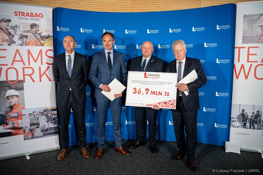 Czterech mężczyzn stoi na tle ścianki z napisem lubelskie. Pozują do zdjęcia. Jeden z nich stojący w środku trzyma w rękach symboliczną kartę z informacją o wysokosci przekazanego dofinansowania