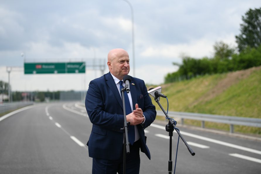 Marszałek Jarosław Stawiarski podczas przemówienia na tle ulicy oraz drogowskazu