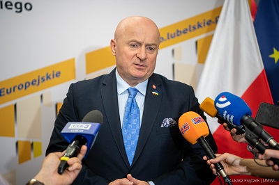 Mężczyzna stoi na tle dwukolorowej ścianki. Ma przed sobą trzy mikrofony i udziela wypowiedzi.