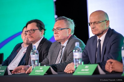 Czterech mężczyzn siedzi za stołem na którym stoją wizytowniki i woda, za nimi ekrany wyświetlające informacje o wydarzeniu sportowym