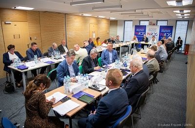 Widok na salę konferencyjną i stoły ustawione w podkowę oraz siedzących przy nich ludzi