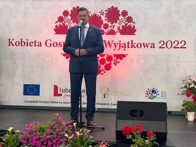 Mężczyzna stoi i przy mikrofonie za nim znajduje się logo wydarzenia kobieta gospodarna wyjątkowa