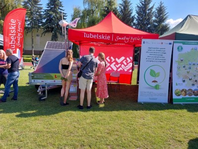 Grupa osób stojących przed stoiskiem Projektu Doradztwa Energetycznego. W tle stoi platforma edukacyjna oze -specjalnie do tego przystosowana przyczepka samochodowa oraz baner reklamowy projektu.