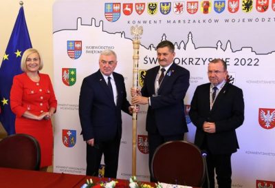 Przekazanie laski marszałkowskiej. Kobieta stoi na tle flagi Unii, 3 mężczyzn stoi na tle ścianki promocyjnej z herbami województw, trzymając Laskę Marszałkowską