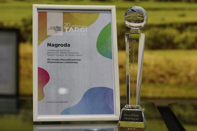 Na stole stoi kryształowy puchar i oprawiony dyplom w tle widać fotografie zielonych pól