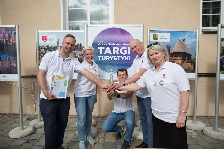 Trzech mężczyzn i dwie kobiety stoją w kręgu i wspólnie trzymają kryształowy puchar, za nimi znajdują się ustawione plakaty z napisem Targi turystyczne