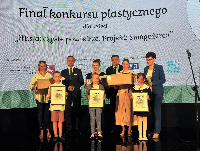 Zwycięzcy w kategorii szkolnej na scenie razem z przedstawicielami organizatorów. W rękach trzymają dyplomy, a ich rodzice nagrody. Za nimi na dużym ekranie nazwa konkursu