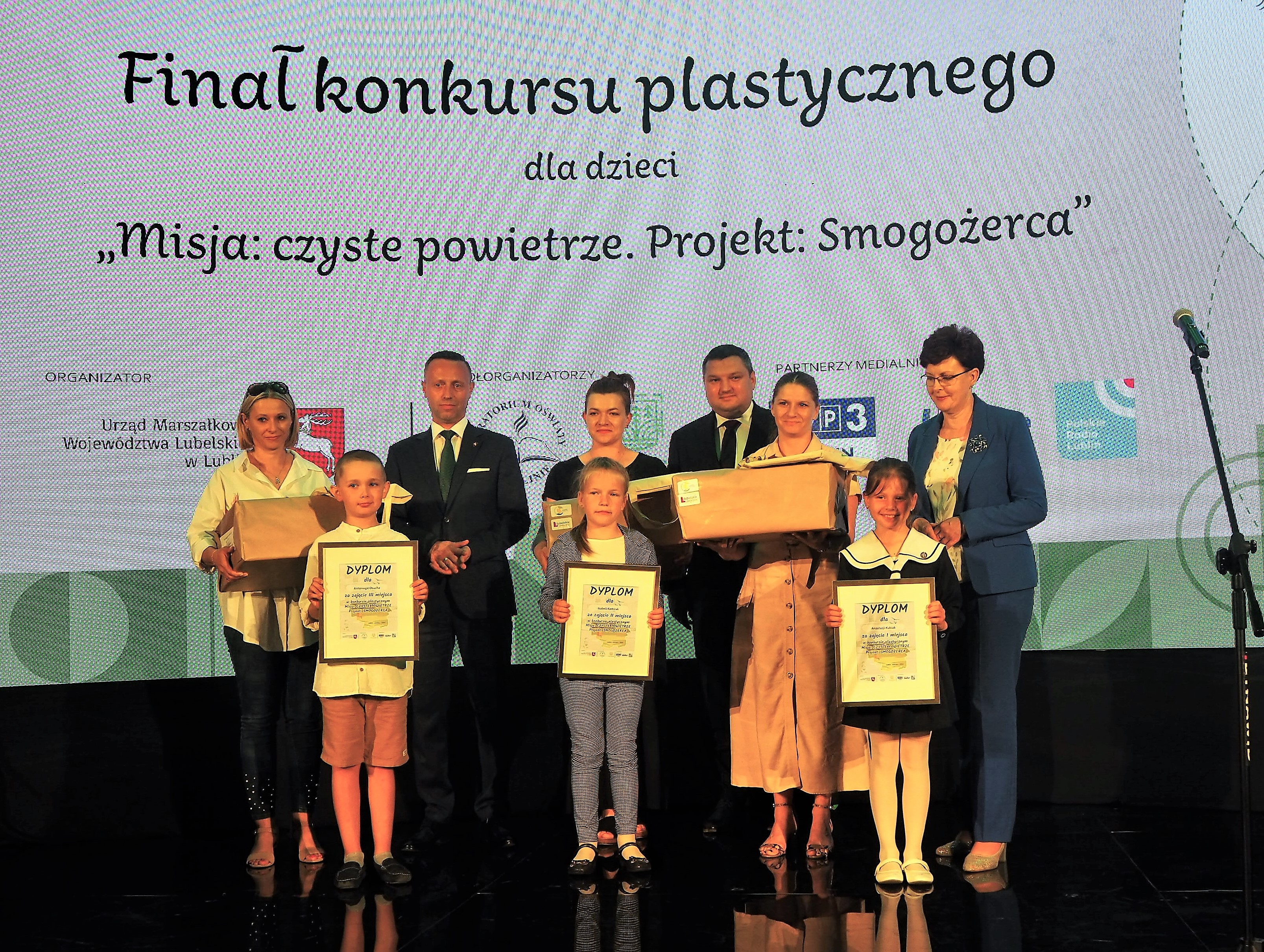 Zwycięzcy w kategorii szkolnej na scenie razem z przedstawicielami organizatorów. W rękach trzymają dyplomy, a ich rodzice nagrody. Za nimi na dużym ekranie nazwa konkursu