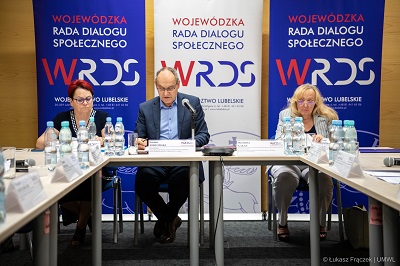 Widok na salę konferencyjną i trzy osoby siedżace przy stole, dwie kobiety i jeden mężczyzna, za nimi znajdują się roll upy promocyjne z napisem WRDS Województwo Lubelskie