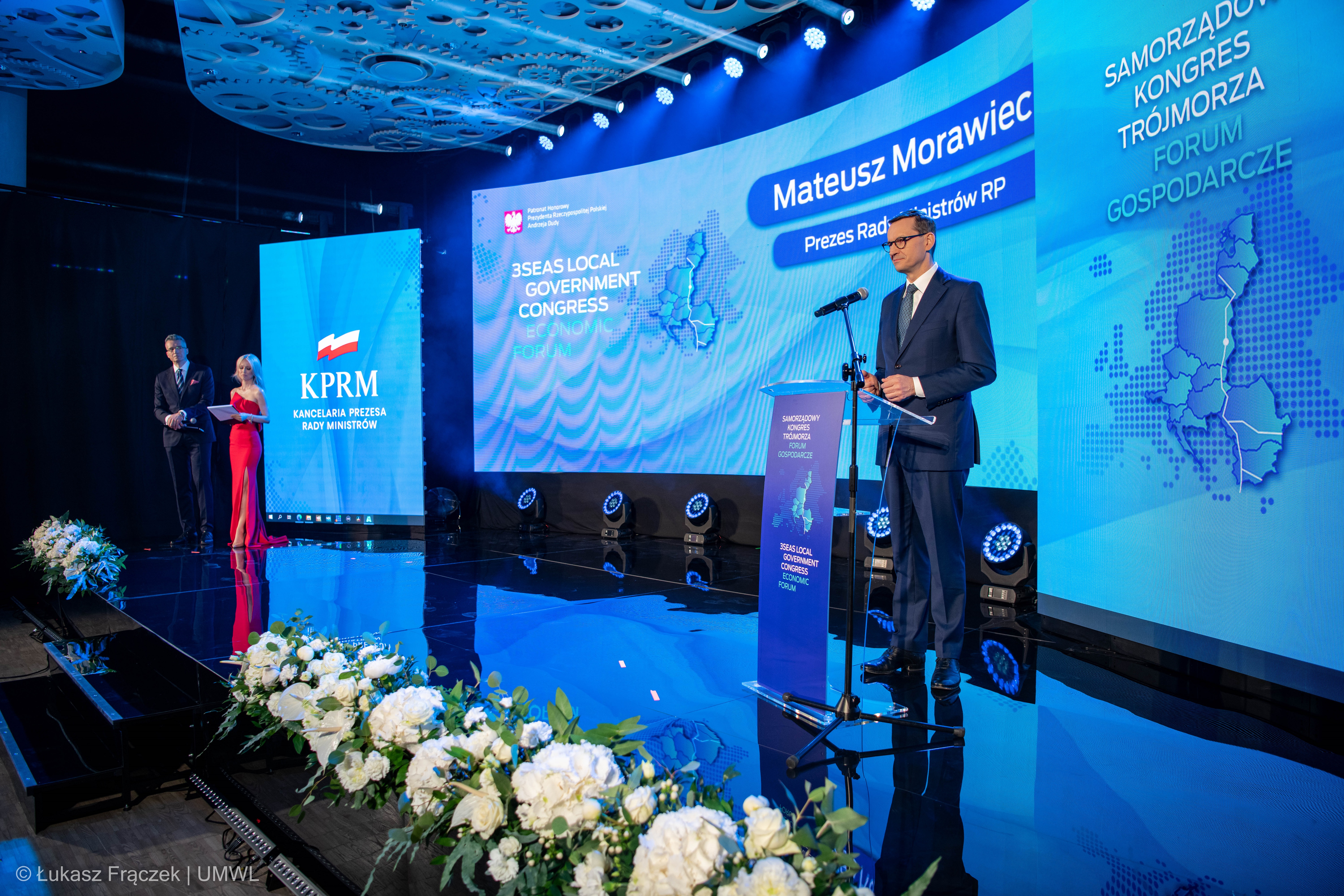 Na scenie przy mikrofonie stoi premier RP. W dali widać dwójkę konferansjerów, którzy stoją i przysłuchują się wypowiedzi premiera. Za nimi znajdują się ekrany z nazwą wydarzenia