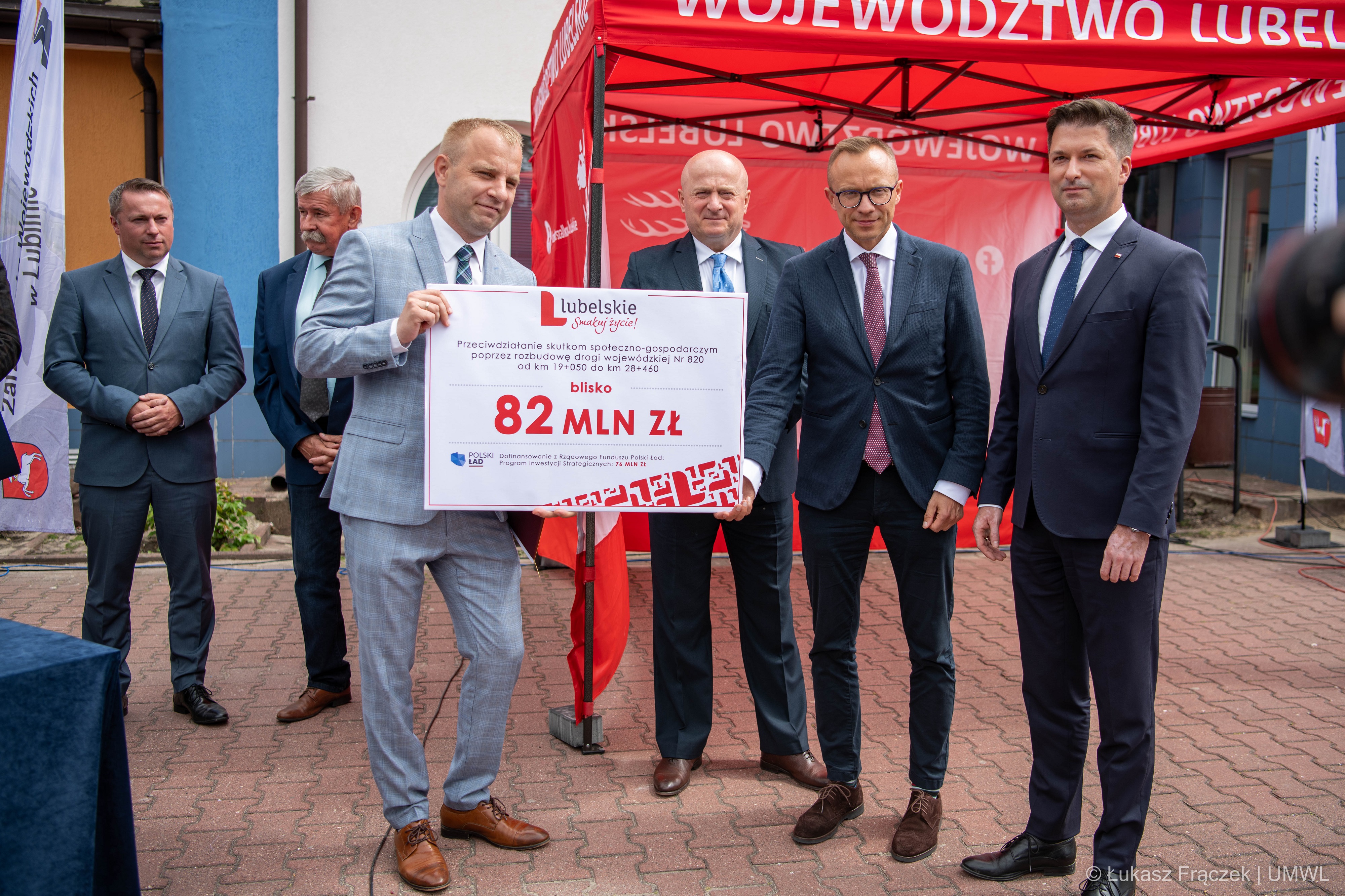 Grupa mężczyzn w garniturach , stoją na zewnątrz i trzymają wspólnie planszę z wypisaną kwotą dotacji na budowę drogi. Za nimi w tle znajduje się namiot promocyjny UMWL