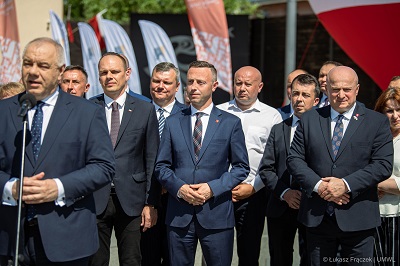 widok na grupę mężczyzn stojących na zewnątrz, jeden z nich po lewej stronie stoi przy mikrofonie i udziela wypowiedzi,