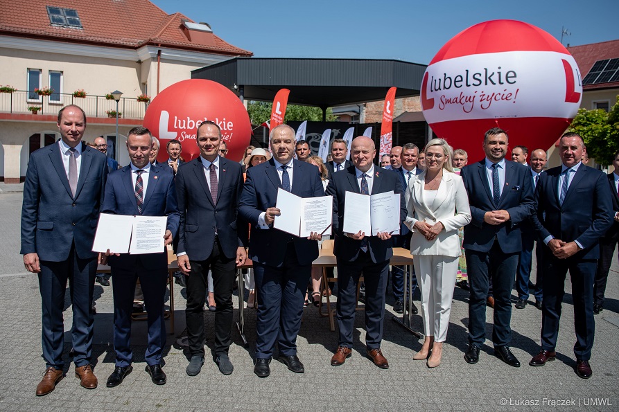 Grupa osób kobiet i mężczyzn stoją na dworze, trzy osoby trzyjmą przed sobą rozłożone teczki i pokazuja podpisane umowy za nimi znajdują się balony promocyjne