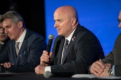 Zbliżenie na profil mężczyzny siedzącego przy stole konferencyjnym. Trzyma w ręce mikrofon i udziela wypowiedzi.