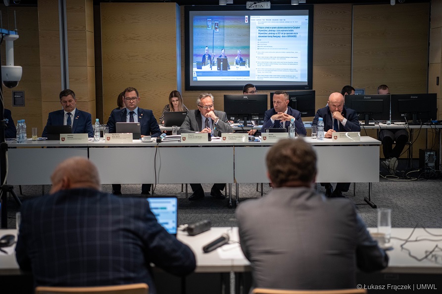 Widok na Członków Zarządu Województwa Lubelskiego, którzy siedzą przy stole konferencyjnym w trakcie trwającej sesji SWL. Za nimi w tle widoczny duzy ekran wyświetlający porzadek obrad spotkania.