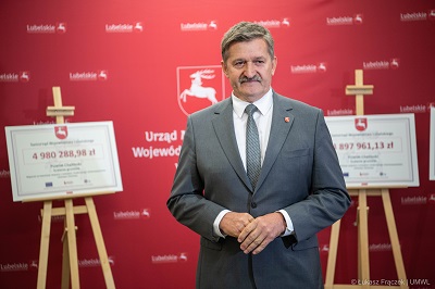 Mężyczna w dojrzałym wieku w garniturze stoi w sali konferencyjnej, za nim na paletach znajdują się tablice z wypisanymi kwotami dotacji dla poszczególnych powiatów